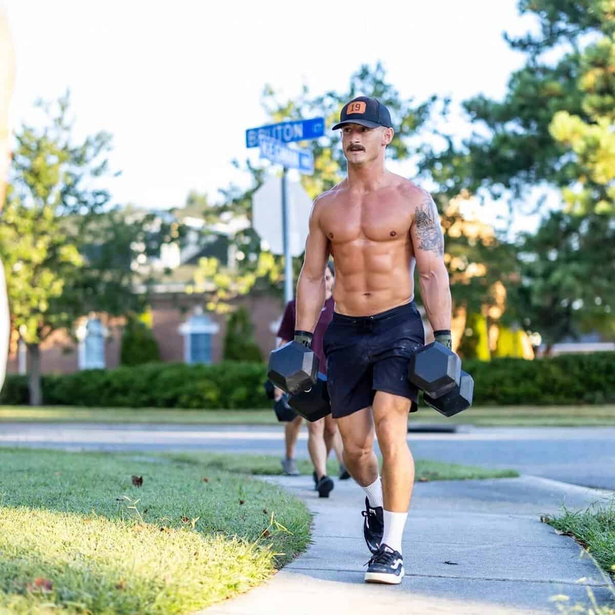 Male athlete walks shirtless with dumbbells on a sidewalk.