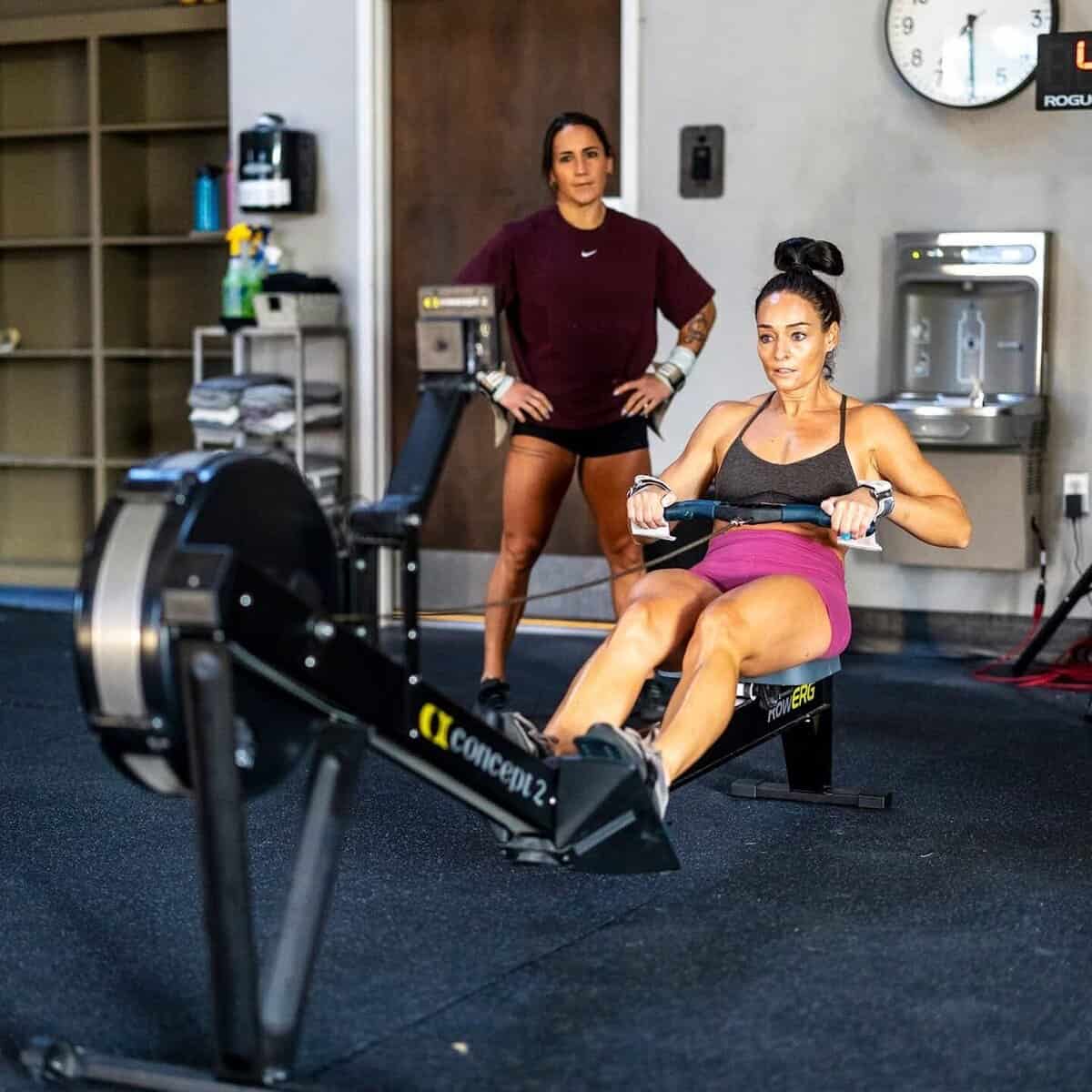 Gym member rows intensely while a coach observes, enhancing fitness tracking.