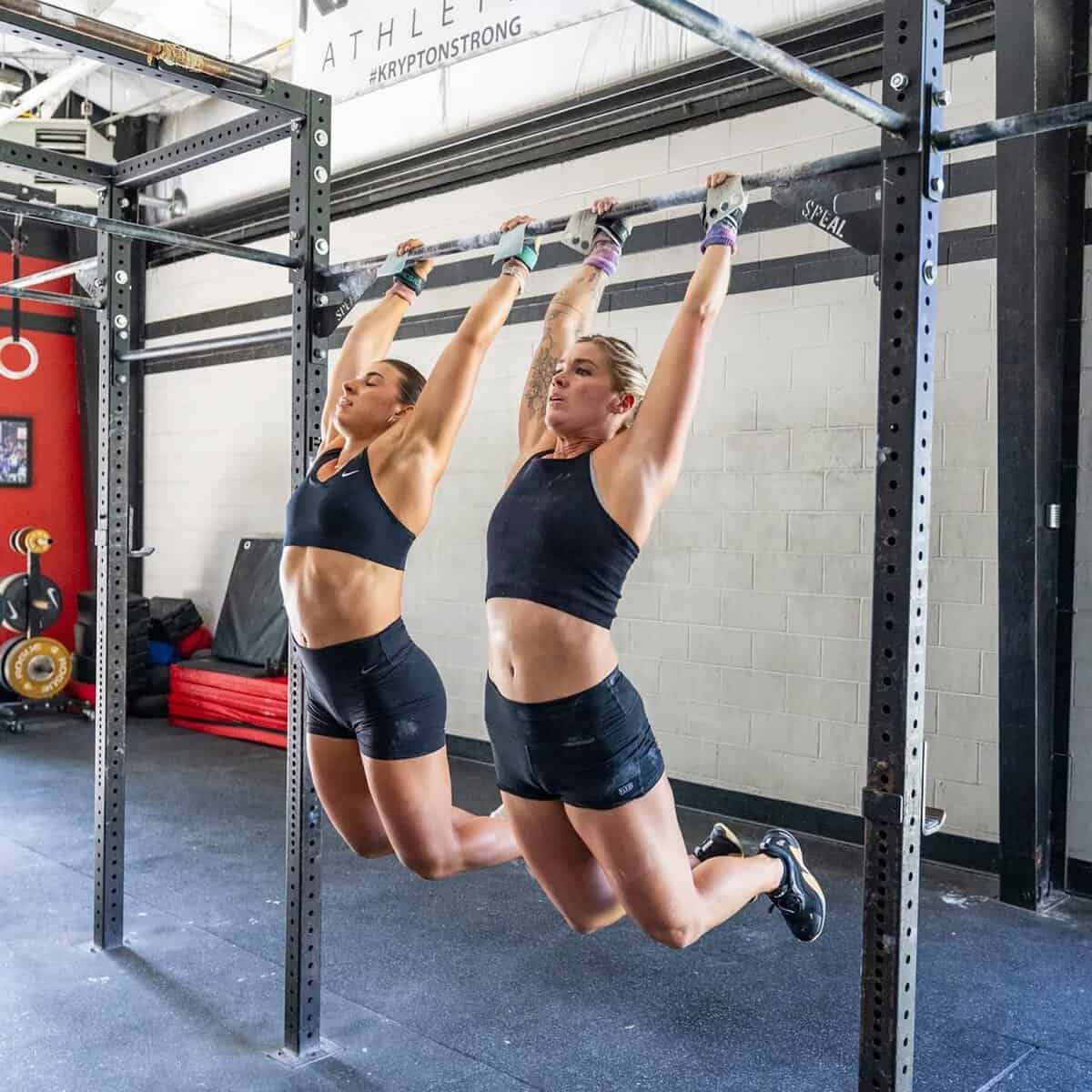 Two athletes hang from a pull-up bar in a gym.