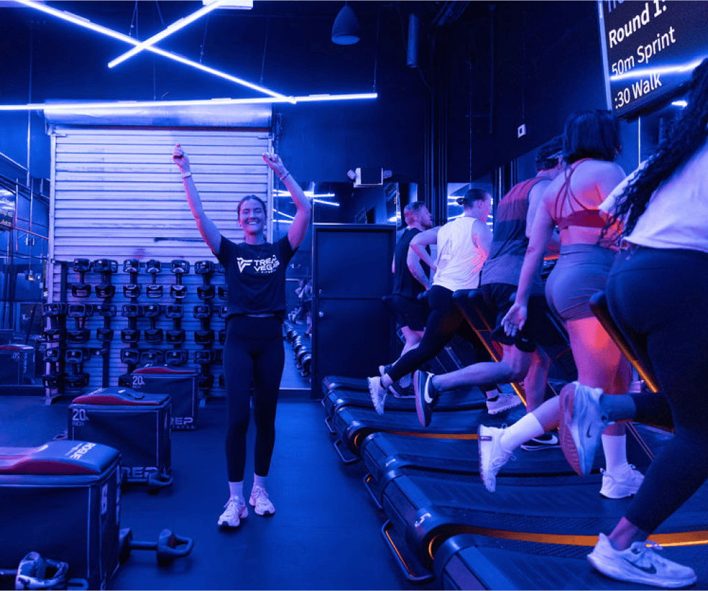 A woman celebrates in a gym with people running on treadmills, showcasing Wodify's fitness tracking.
