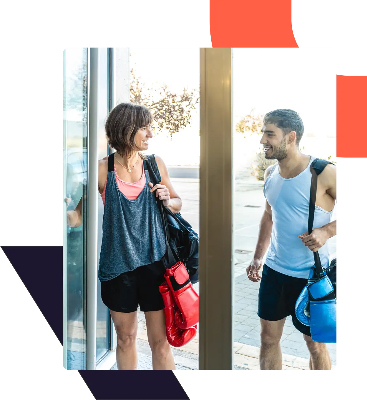 Two people with gym bags and boxing gloves smiling, entering a gym.