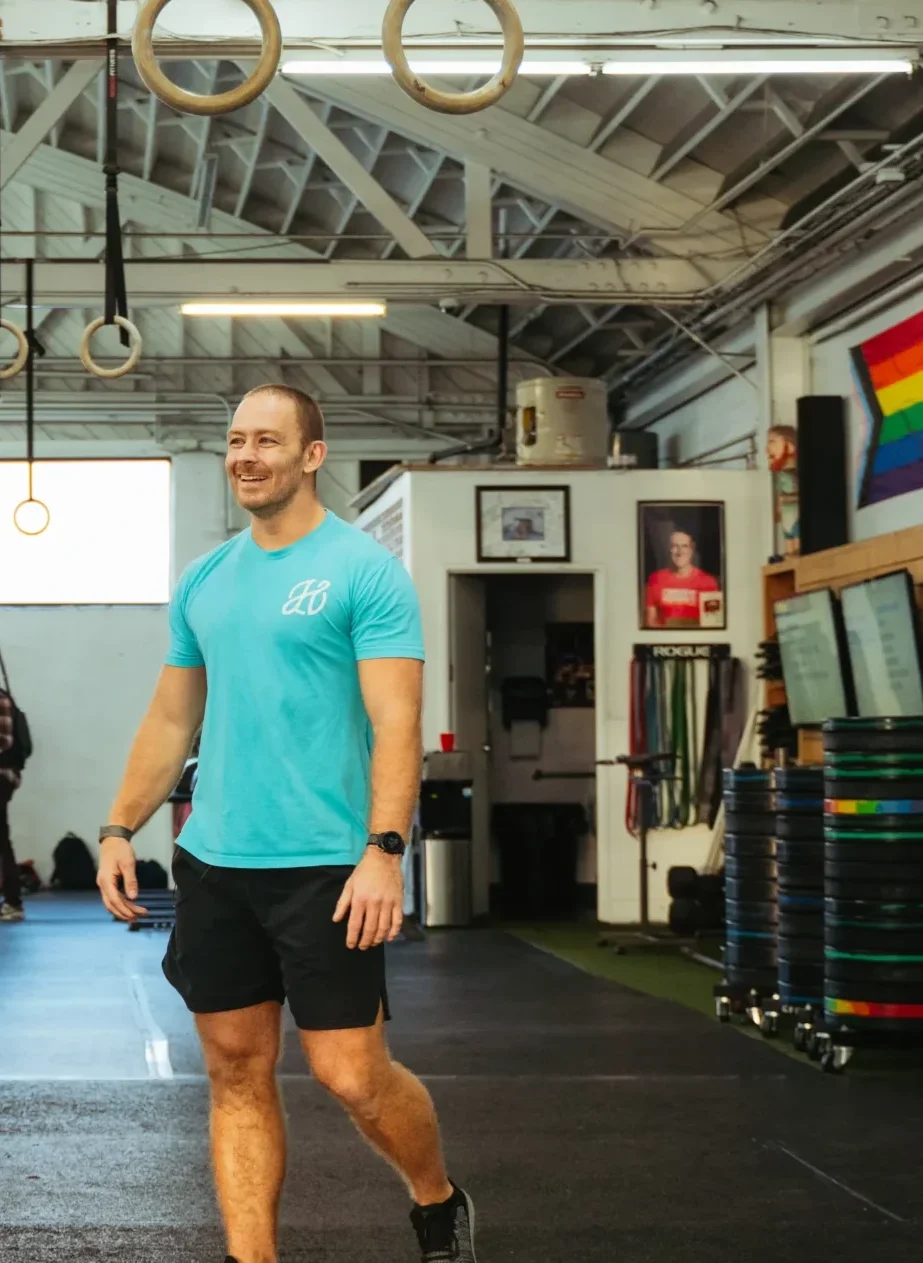 Craig Wayne in a gym, smiling while walking past gymnastic rings and weights.