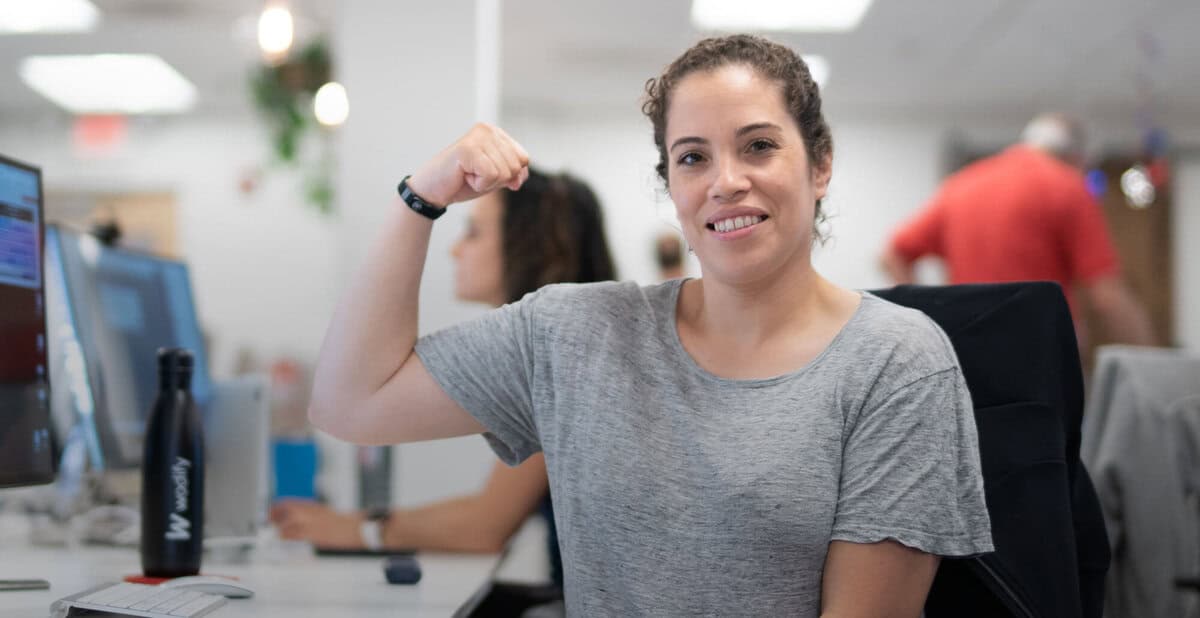 A smiling Wodify employee flexes her bicep in a bright, modern office.