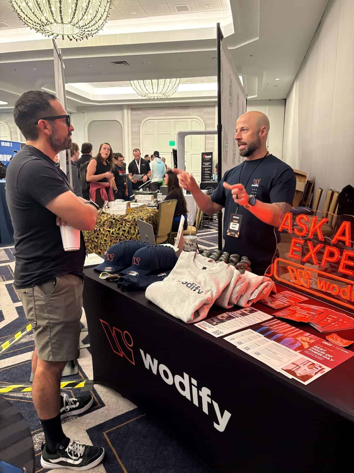A Wodify representative talks to a man at a branded booth in a convention hall.