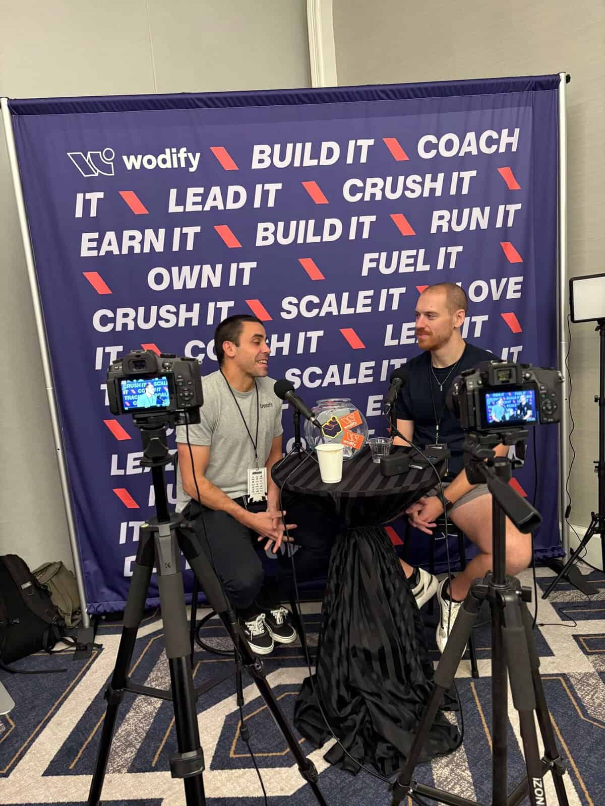 Two guys podcasting in front of a Wodify branded backdrop with two cameras filming.