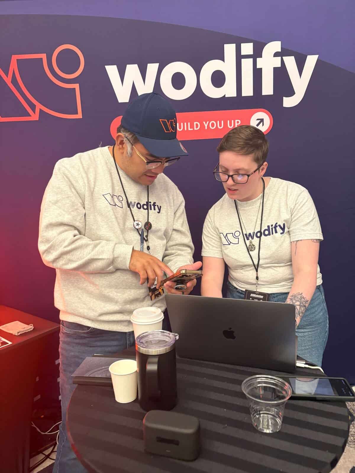Wodify employees discuss content on a phone and laptop at a branded booth.