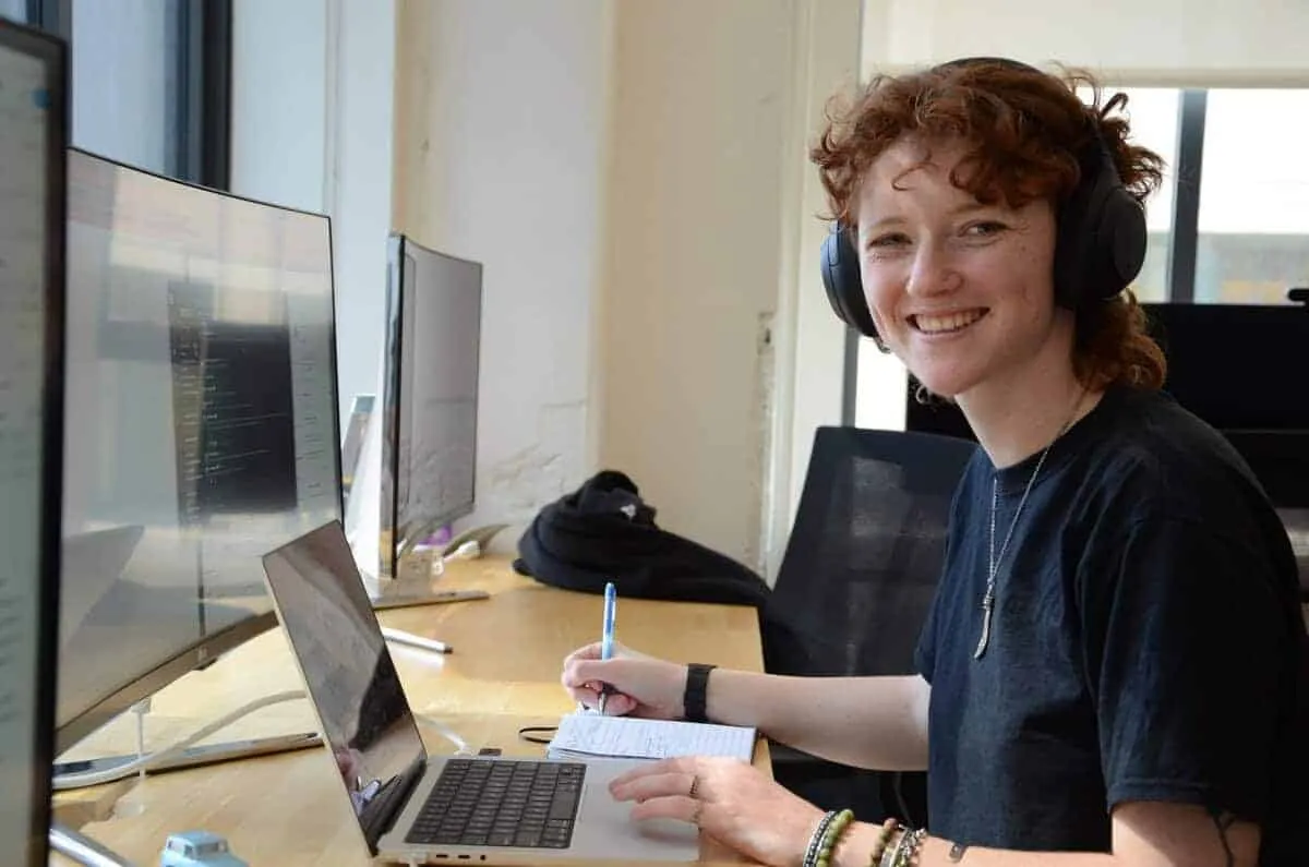 Wodify team member smiling, wearing headphones, writing notes at a laptop in an office.