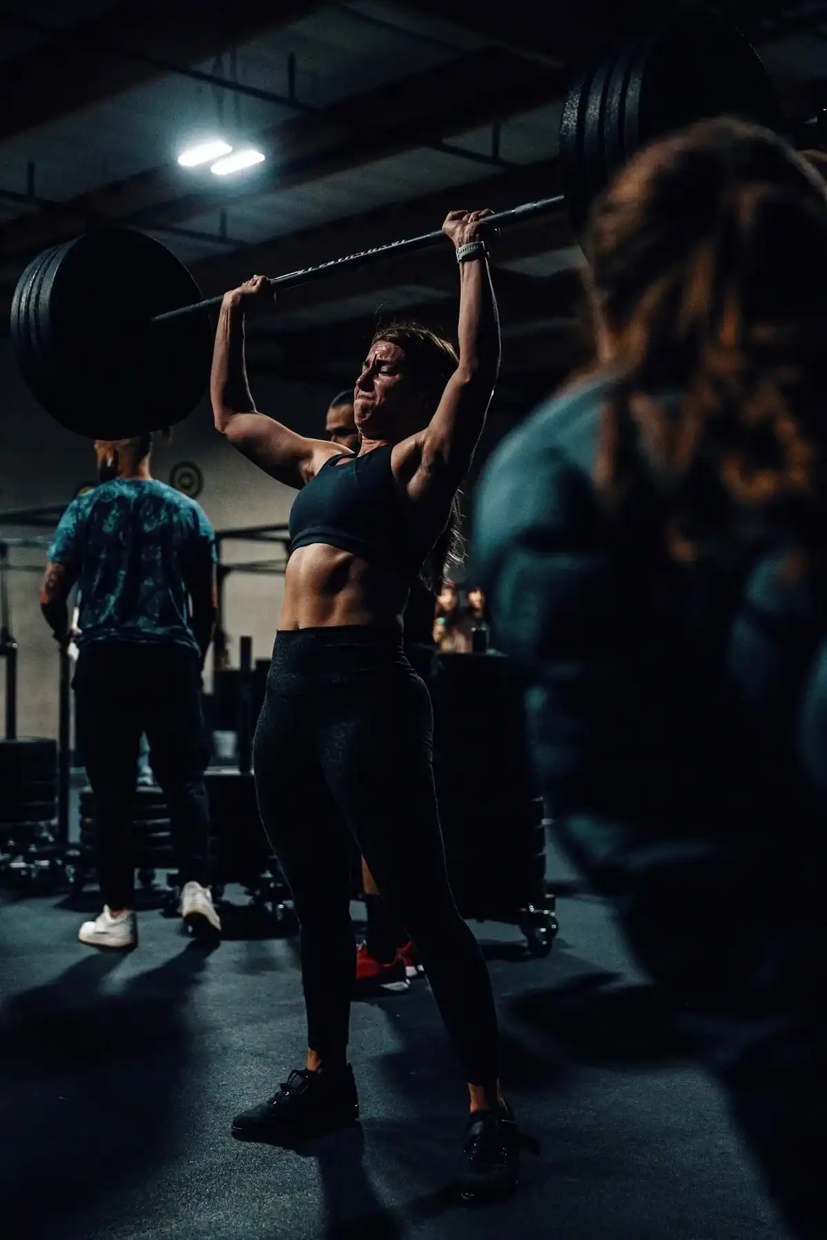 Wodify athlete lifting heavy barbell overhead in a dark gym, focused on strength.